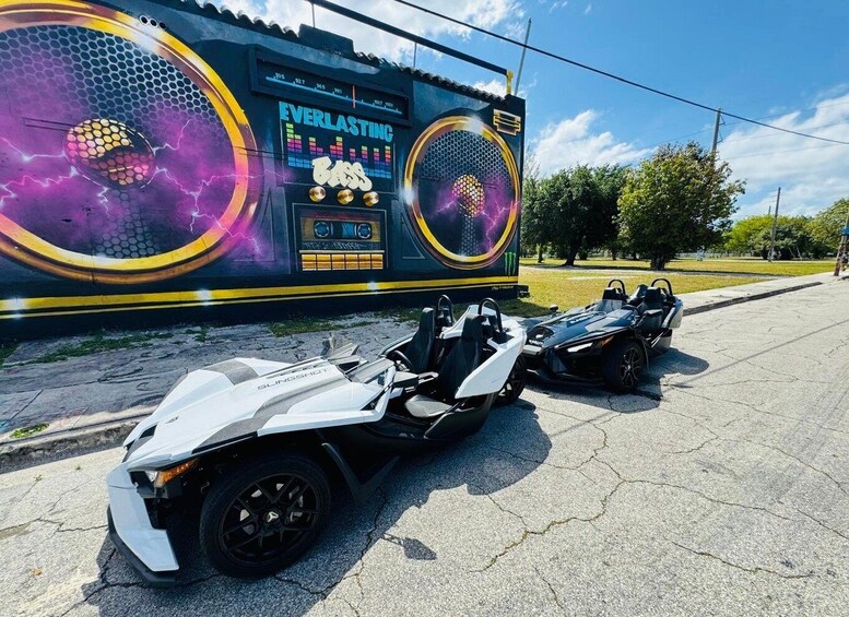 Two racing cars parked in front of a large, colorful billboard with the text 'EVERLASTING' and 'Bass' on it. 2