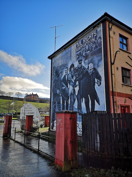 A mural on a building depicting a historical event, a sign reading 'You are now entering Free Derry,' and a red gate with a fence. 1