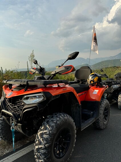 A red 4x4 ATV with a flag, parked on a road with mountains in the background. 3