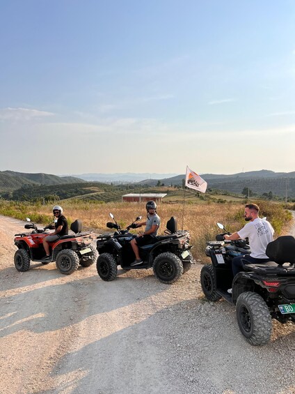 Three people on ATVs on a gravel path with a flag, hills, and a building in the background. 1