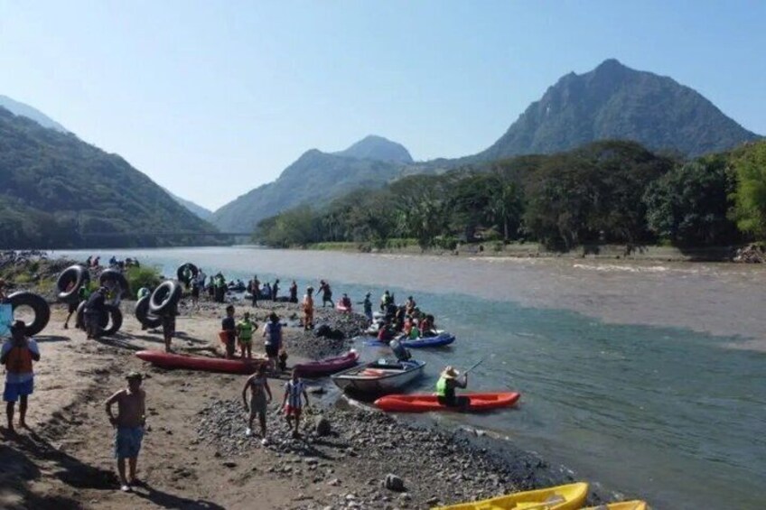 A group of people on a riverbank with canoes and kayaks, surrounded by mountains. 3