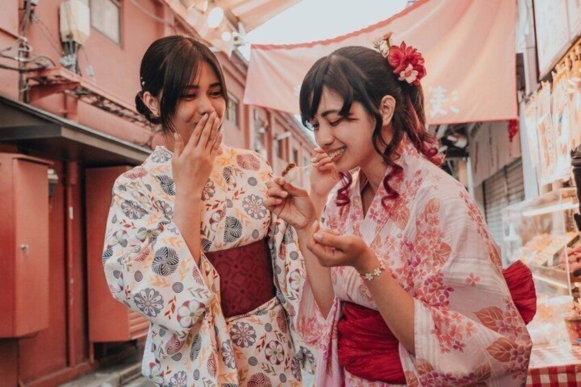 Two individuals in traditional attire, one holding a fan, standing in an outdoor market with red buildings and a pink canopy. 3