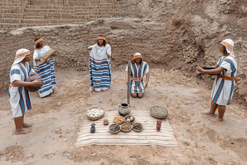 A group of people in traditional attire around a table with various items, including pottery and food, in an outdoor setting with a brick structure in the background. 1