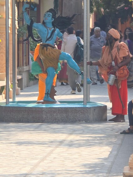 A statue of a blue-skinned figure with a raised arm, a person in orange attire, and a crowd in the background. 1