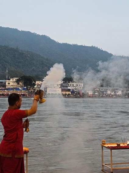 A person performing a ritual by the water, with smoke rising and a temple in the background. 5