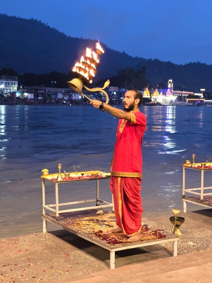 A person in a red robe performing a ritual by the water with a fire torch, surrounded by a temple and a river. 4