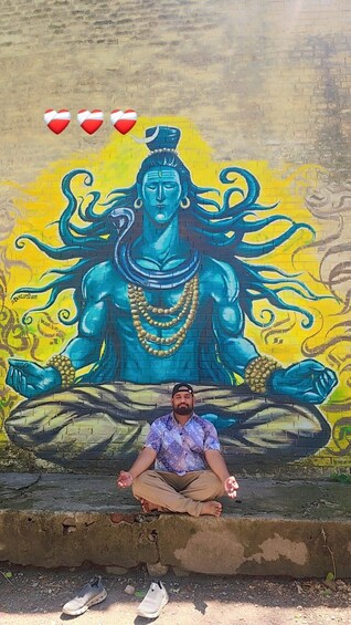 A man sitting in a meditative pose in front of a mural of a deity. 2