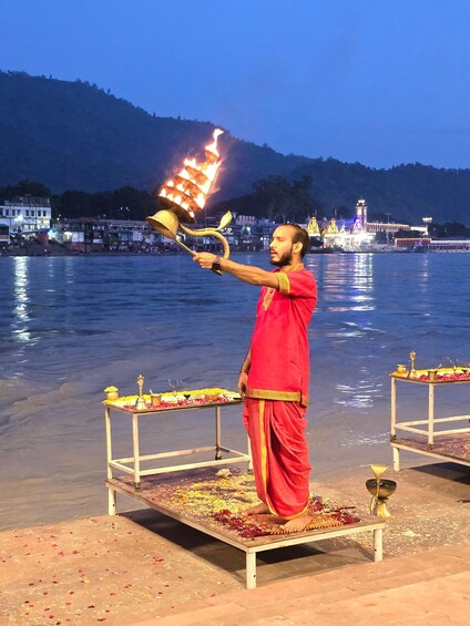 A person performing a ritual by the water with a flaming lamp, surrounded by ritualistic items and a temple in the background. 3