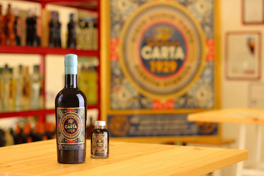 A bottle of Carta 1929 and a small bottle on a wooden table with a Carta 1929 poster in the background. 1