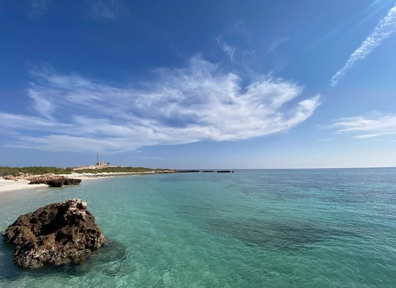 A clear turquoise sea, a rocky outcrop in the foreground, and a distant shoreline with sparse vegetation and a structure. 5