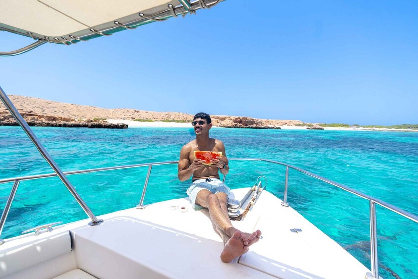 A person on a boat in clear turquoise waters, holding a slice of watermelon. 1