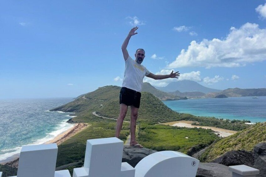 A person standing on a cliff with arms raised, overlooking a coastal landscape with hills and the ocean. 4