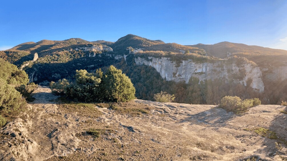 A rugged mountain landscape with a prominent cliff face, sparse vegetation, and a clear sky. 1