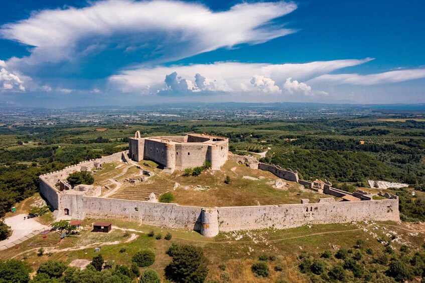 A historic stone fortress with a central tower, surrounded by a fortified wall and lush greenery. 1