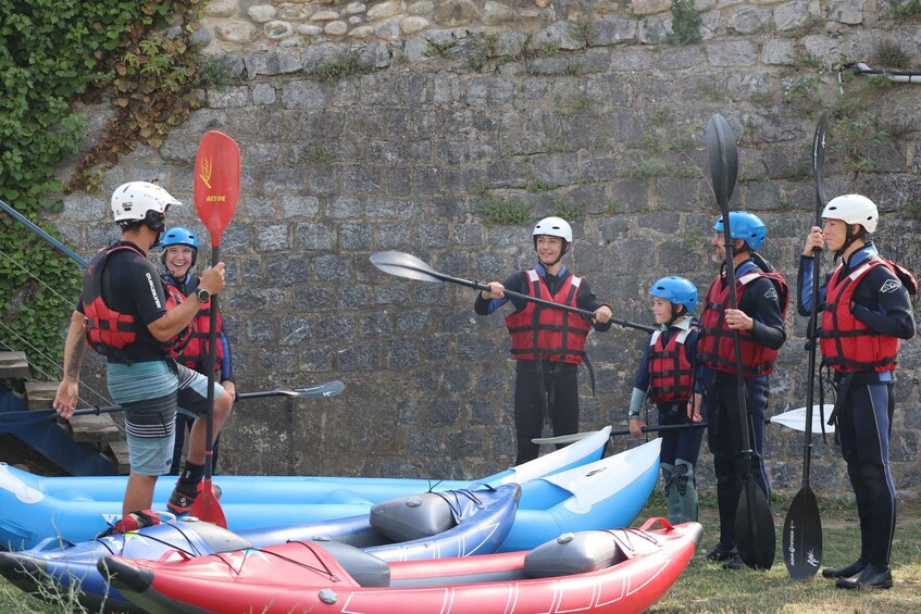 A group of people in life jackets and helmets standing around kayaks and paddles. 2