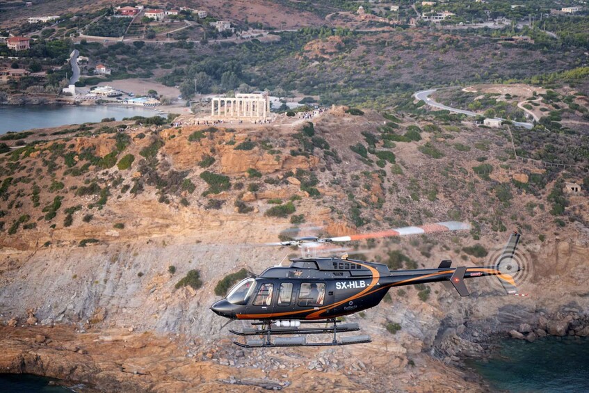A helicopter with 'SX-HLB' and 'Bellatrix' on its side flying over a coastal area with rocky terrain and a building in the background. 1