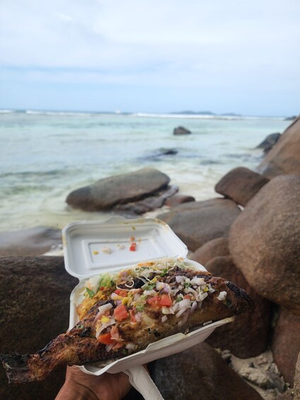 A grilled fish on a white plate with a side of coleslaw and diced tomatoes, held over a rocky beach with the ocean in the background. 5