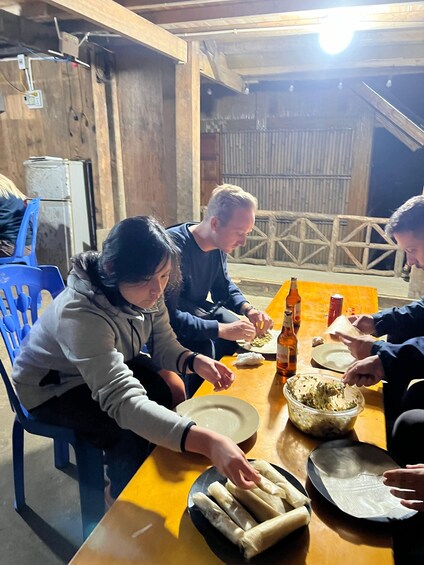Three people preparing food at a wooden table with a bowl of chopped vegetables, a plate of rolled dough, and bottles of beer. 3
