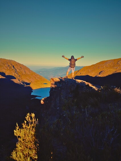 A person standing on a cliff edge with arms outstretched, overlooking a valley and a body of water. 5
