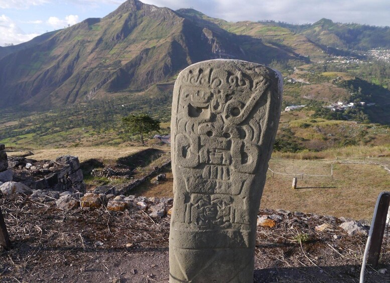 A stone monument with carvings, set against a mountainous landscape with a village in the distance. 5
