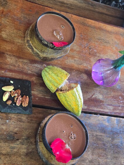 Two wooden cups with a brown liquid, a halved green nut, a purple flower, and some nuts on a wooden surface. 1