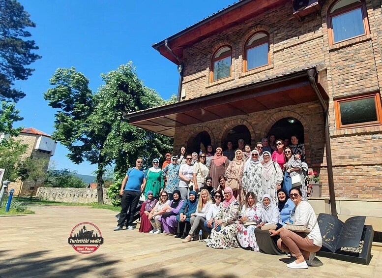 A group of people posing in front of a stone building with arched windows and a tiled roof. 1