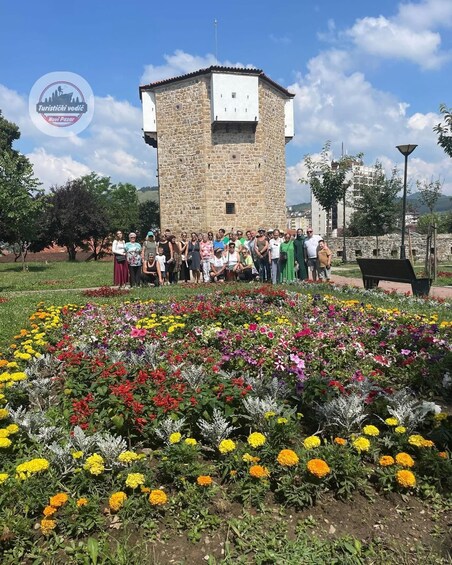 A group of people standing in front of a stone tower with a flower garden in the foreground. 3