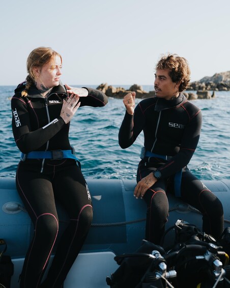 Two people in wetsuits sitting on a boat with the ocean and rocks in the background. 4