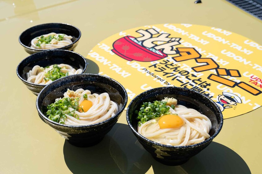 Three bowls of ramen with green onions and a fried egg on a yellow surface with a 'UDON-TAXI' logo. 5