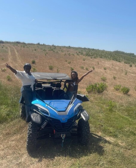 Two people standing beside a blue off-road vehicle on a grassy hill with a clear sky. 1