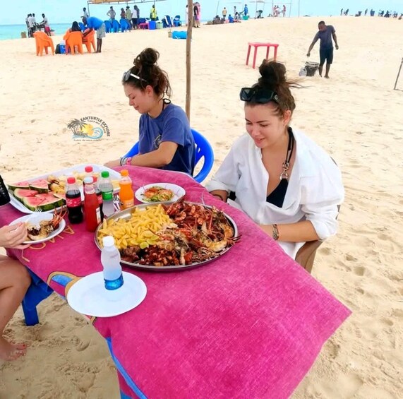 A beachside dining setup with a table covered in a pink cloth, featuring a seafood platter, drinks, and a watermelon slice. 2