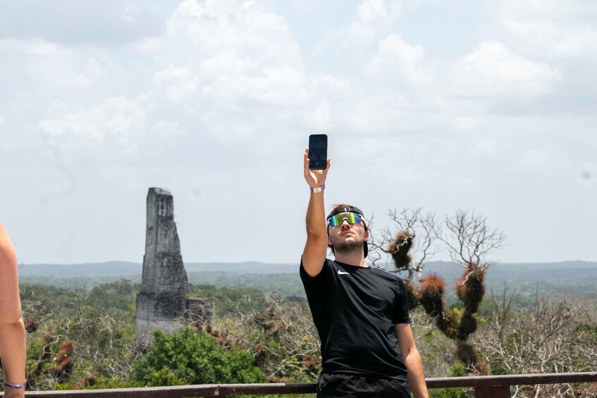 A person taking a photo with a smartphone, a large ancient structure in the background, and a landscape with sparse vegetation. 2