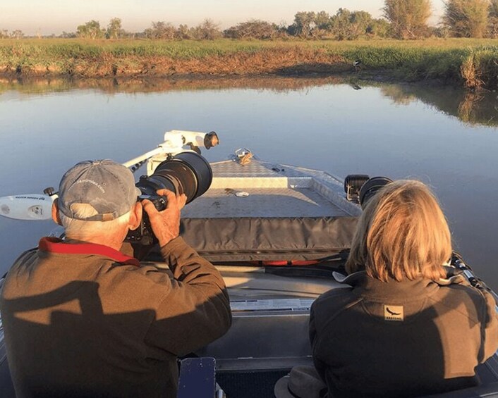 Two people on a boat, one using a camera, with a calm river and greenery in the background. 4
