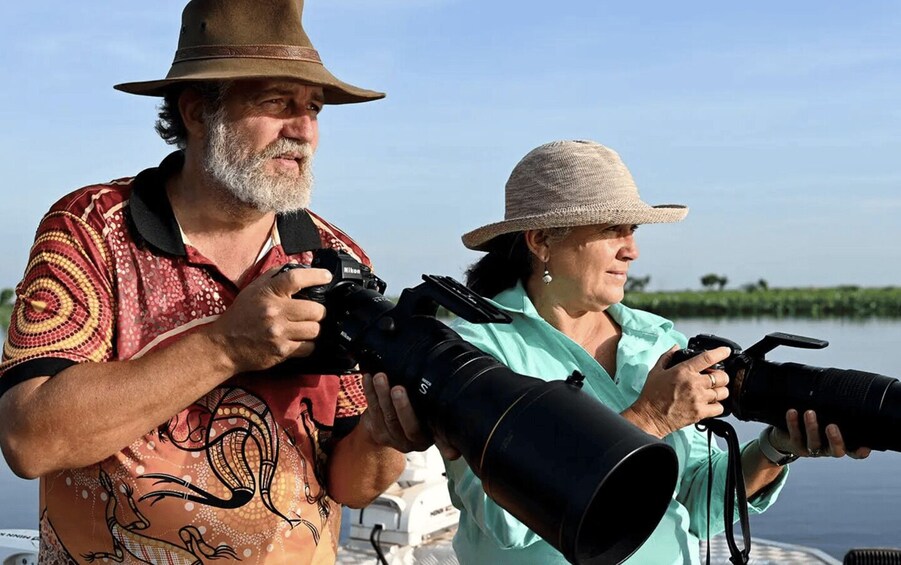Two people with cameras, one holding a Nikon and the other a Canon, on a boat with water and greenery in the background. 3