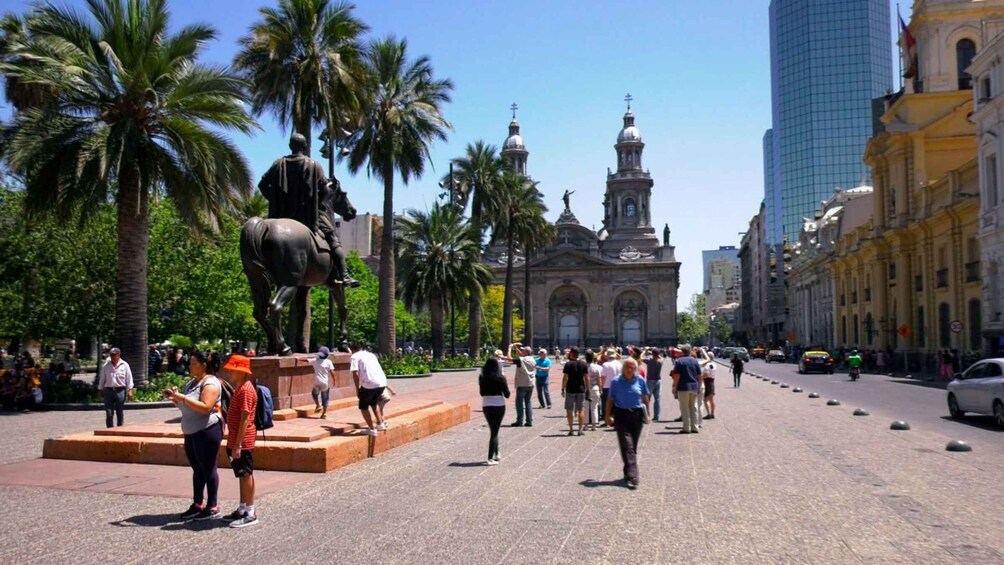 A plaza with a statue, palm trees, a church with domes, and modern buildings. 1