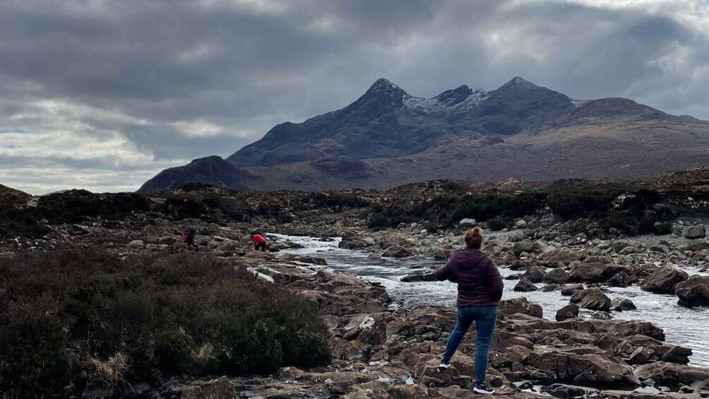 A rocky riverbed with a person in a purple jacket standing on rocks, a person in a red jacket walking in the distance, and mountains in the background. 5