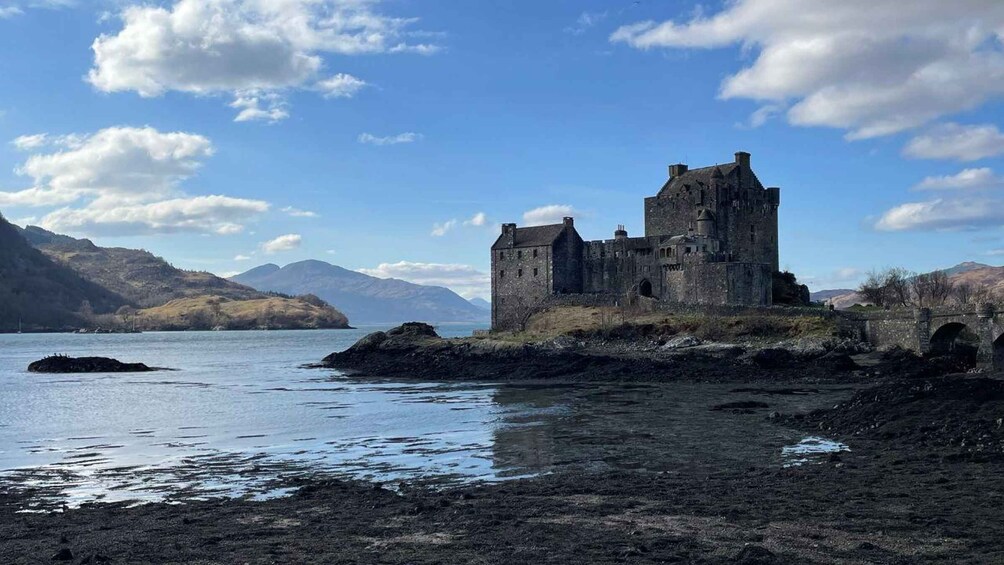 A historic castle on a rocky shoreline with a lake and mountains in the background. 1
