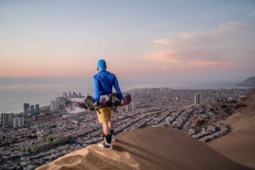 A person standing on a sand dune overlooking a cityscape with buildings and a body of water. 2