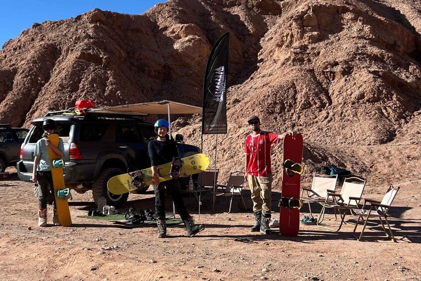 A group of people with snowboards, a vehicle, and a flag in a rocky outdoor setting. 3