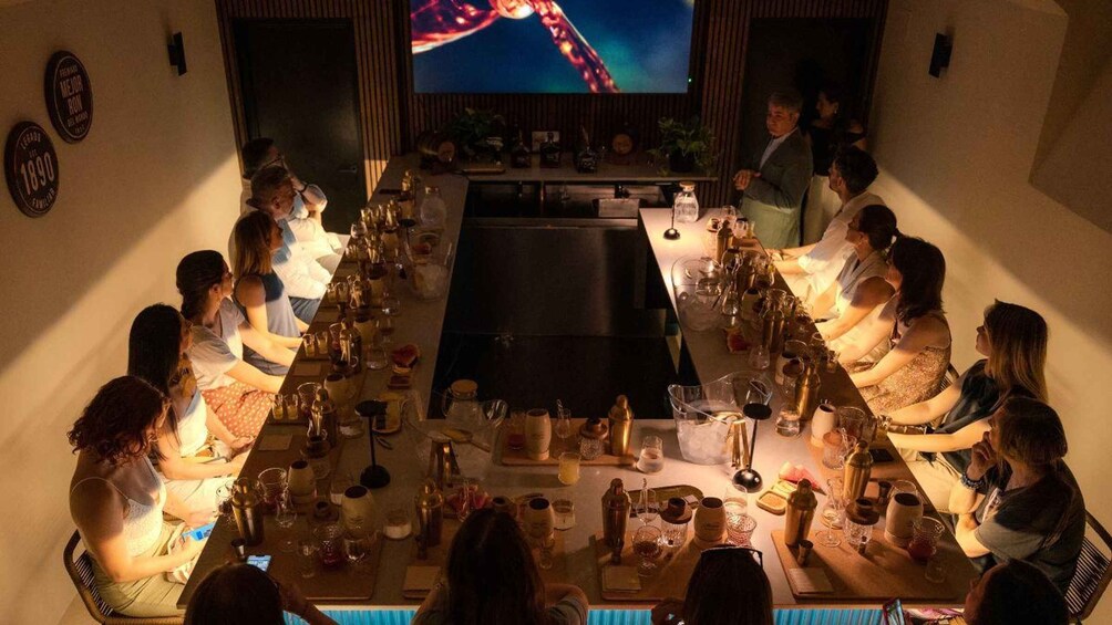 A group of people seated around a long table in a dimly lit room with a television screen displaying a colorful image in the background. 2