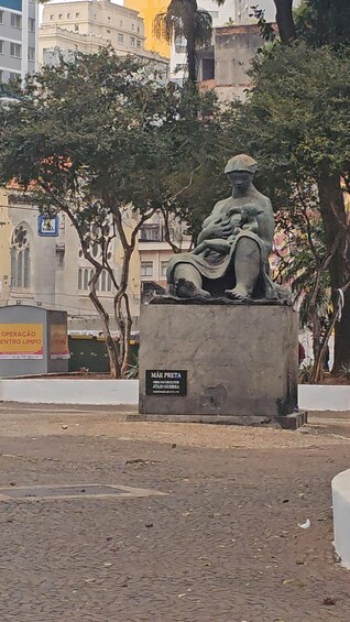 A statue of a seated figure with a plaque reading 'MAE PRETA' in a park with trees and buildings in the background. 3