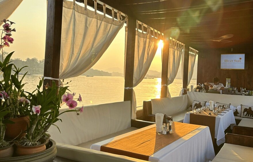 A restaurant with a view of the sunset over water, featuring white curtains, wooden furniture, and a sign reading 'River Sun.' 5