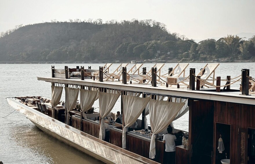 A boat with a deck area featuring wooden furniture and white curtains, set against a backdrop of a calm river and a forested hill. 2