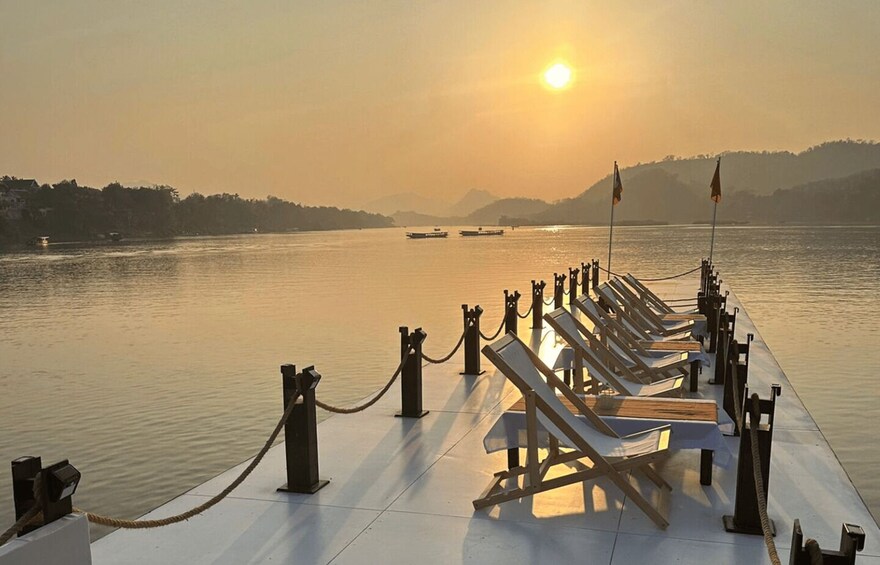 A riverside pier with lounge chairs and a rope barrier during sunset. 1
