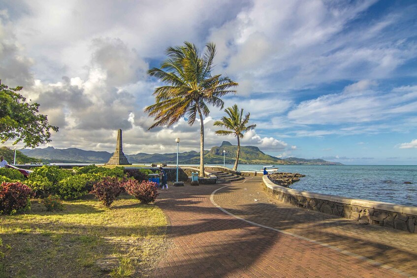 A coastal walkway with palm trees, a stone monument, and a view of the sea and mountains. 3