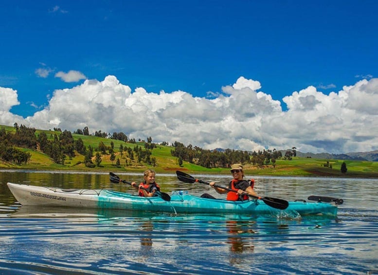 Two people kayaking on a calm lake with a clear sky and green hills in the background. 1