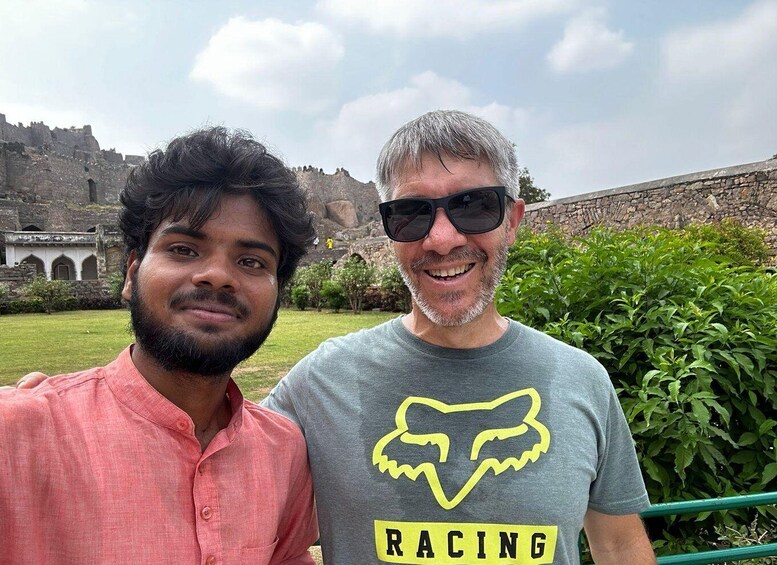 Two individuals in front of a historical fort with greenery and a stone wall. 3