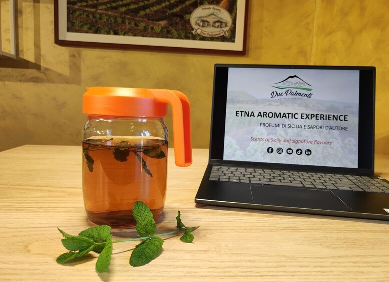 A laptop displaying a website for ETNA Aromatic Experience, a jar of tea with mint leaves, and a framed picture on the wall. 3