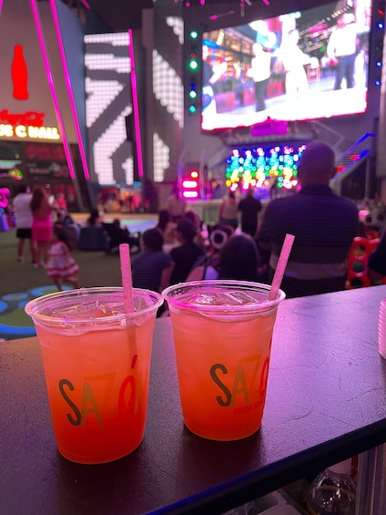 Two drinks with straws labeled 'SA' on a table, with a neon-lit background and a crowd in the distance. 3