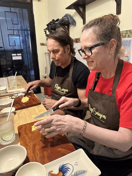 Two individuals wearing aprons with 'Nyo Saigon' branding, engaged in food preparation on a wooden cutting board. 5